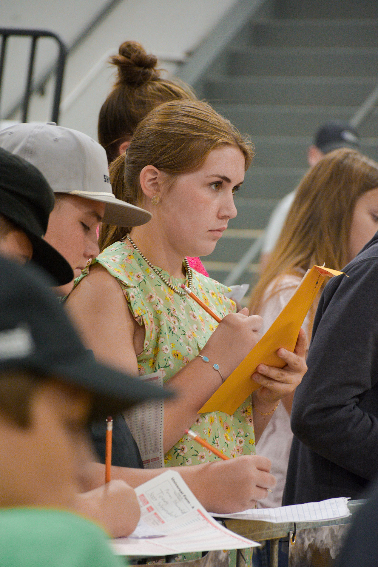 Group of people standing in line with pencils and papers, including one individual writing on an orange envelope near a staircase and railings.