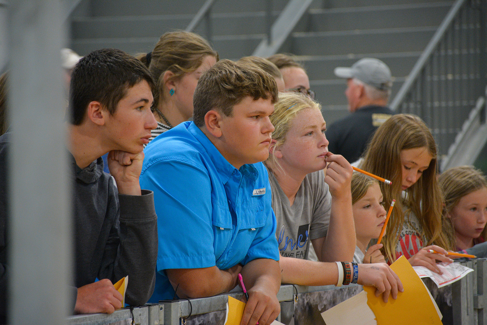 Group of individuals standing behind a metal barrier in an indoor arena, holding papers and pencils during a livestock judging event.