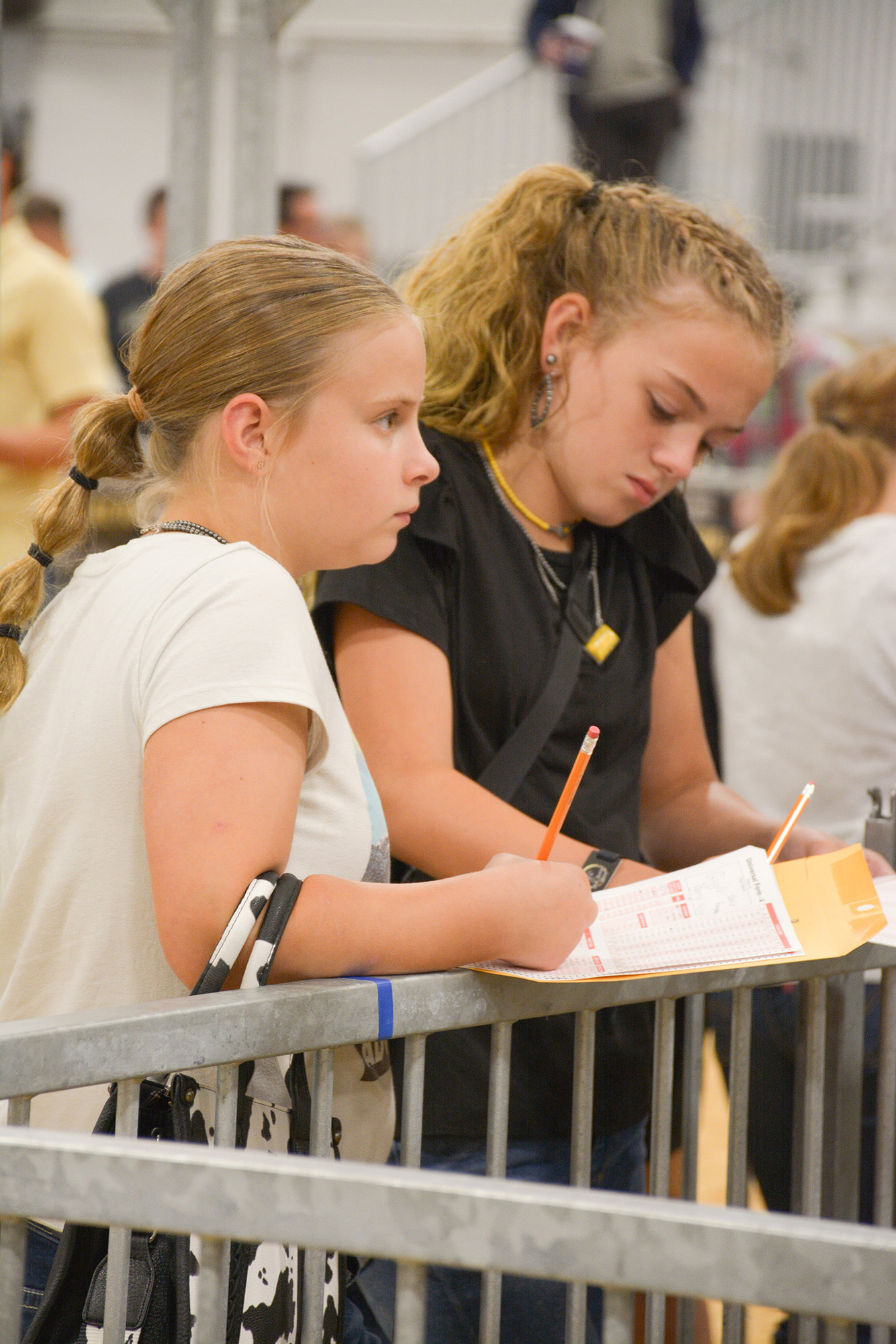 Two individuals writing on forms with pencils in an indoor setting, surrounded by other participants.