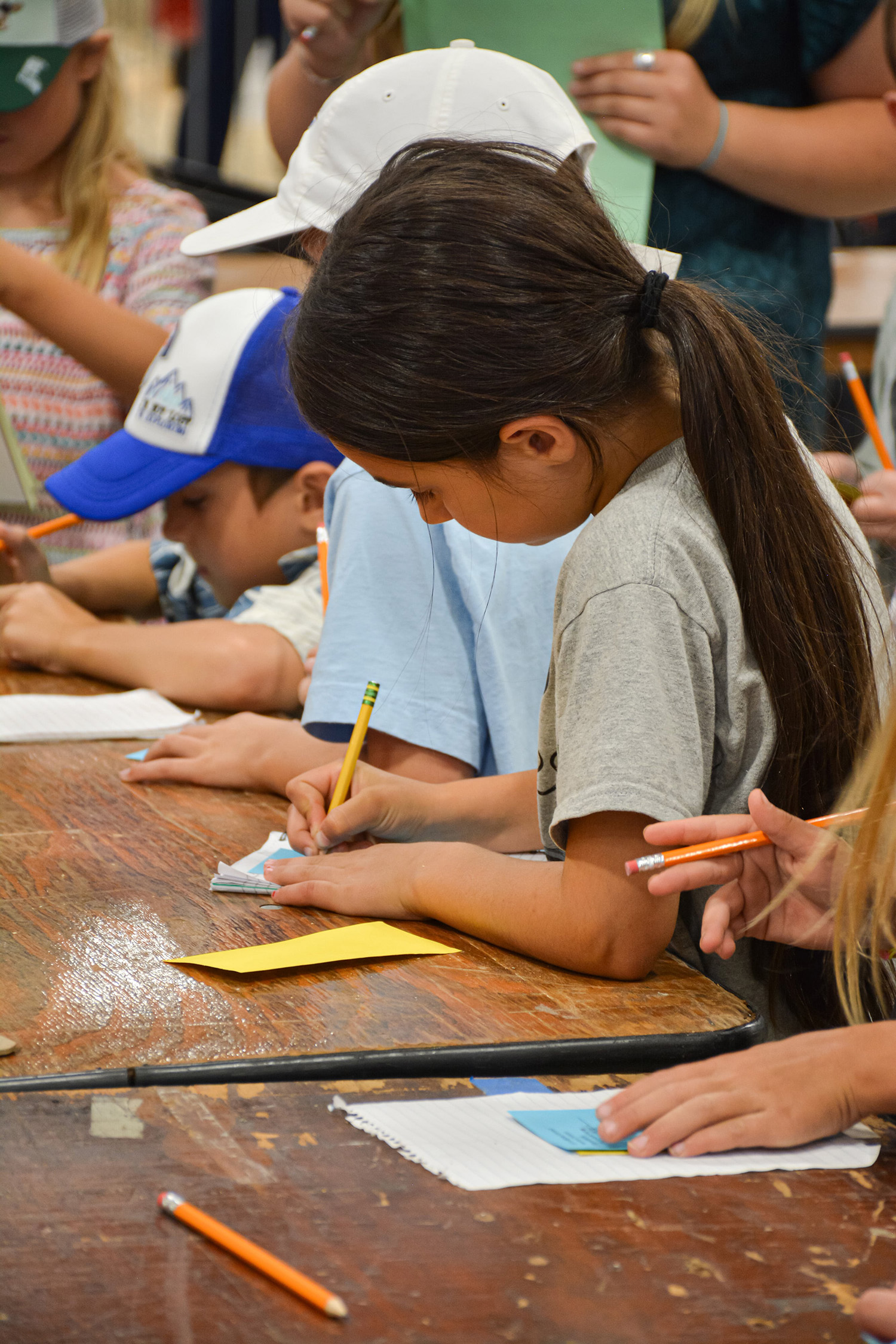 Children sitting at a wooden table, writing or drawing on colored papers with pencils during an indoor activity.