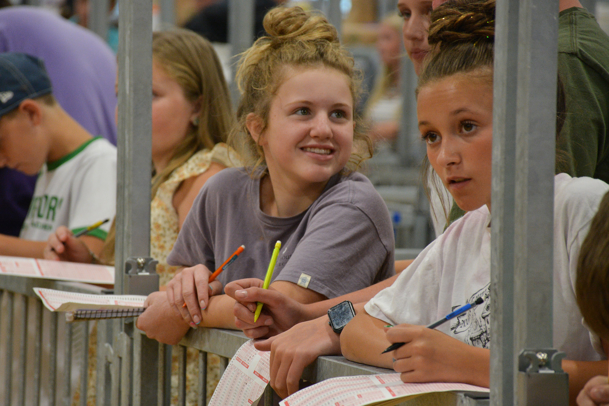 Participants holding pencils and filling out answer sheets in an indoor area with metal railings.