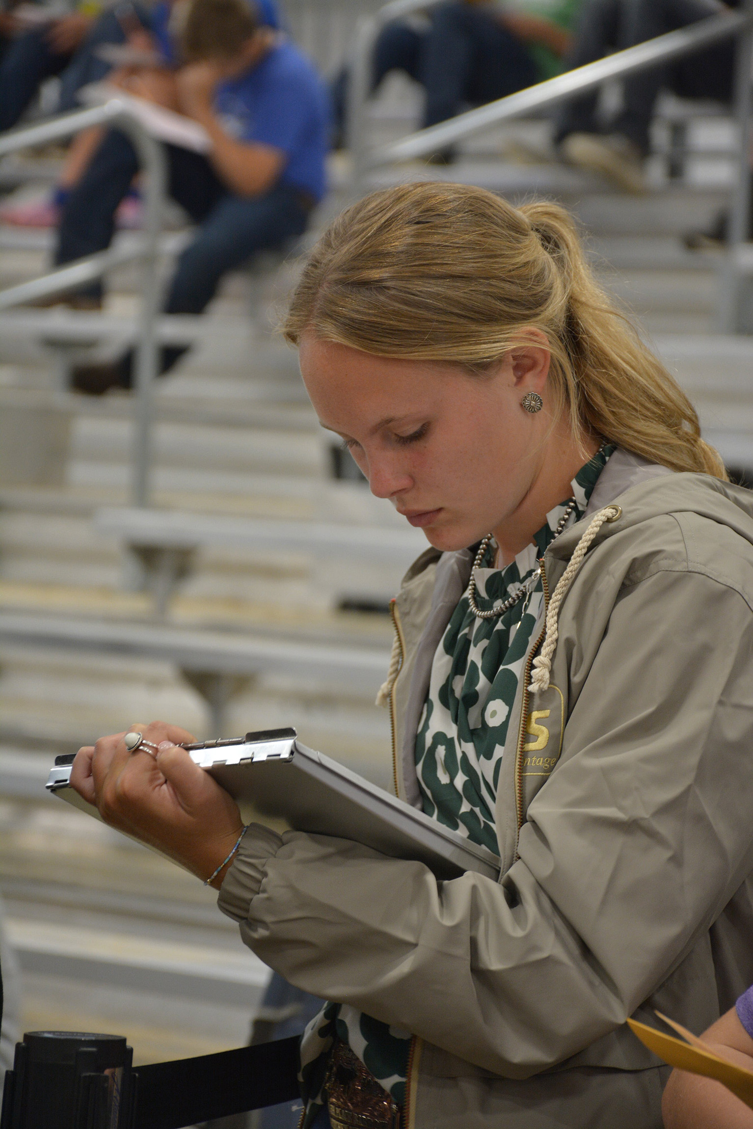 Person wearing a jacket, writing on a clipboard in front of bleachers with seated spectators.