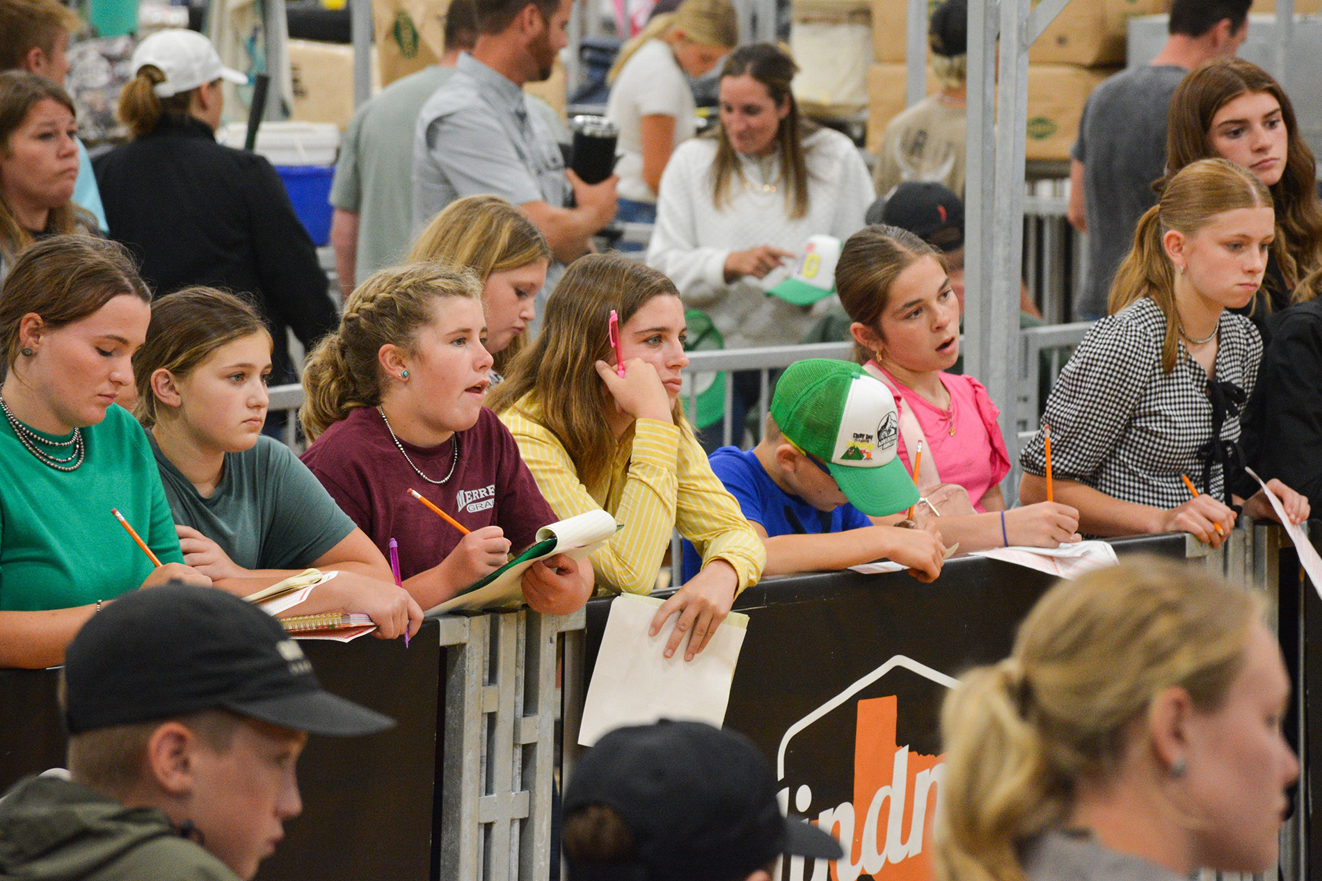 Children and teenagers standing behind a barrier, holding notebooks and pencils while participating in an event.
