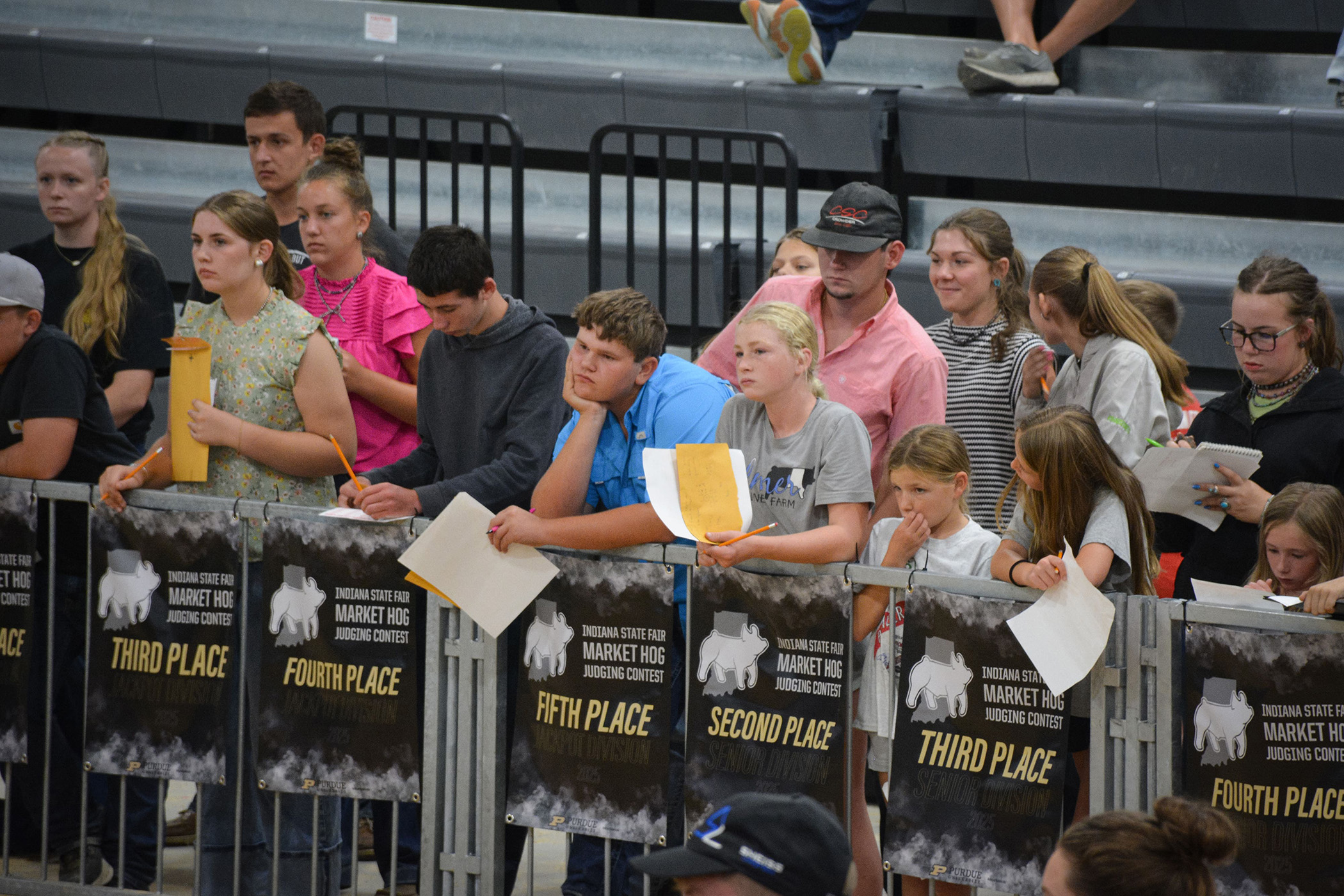 Group of people standing behind a metal barrier with banners displaying placements for the Indiana State Fair Market Hog Judging Contest.