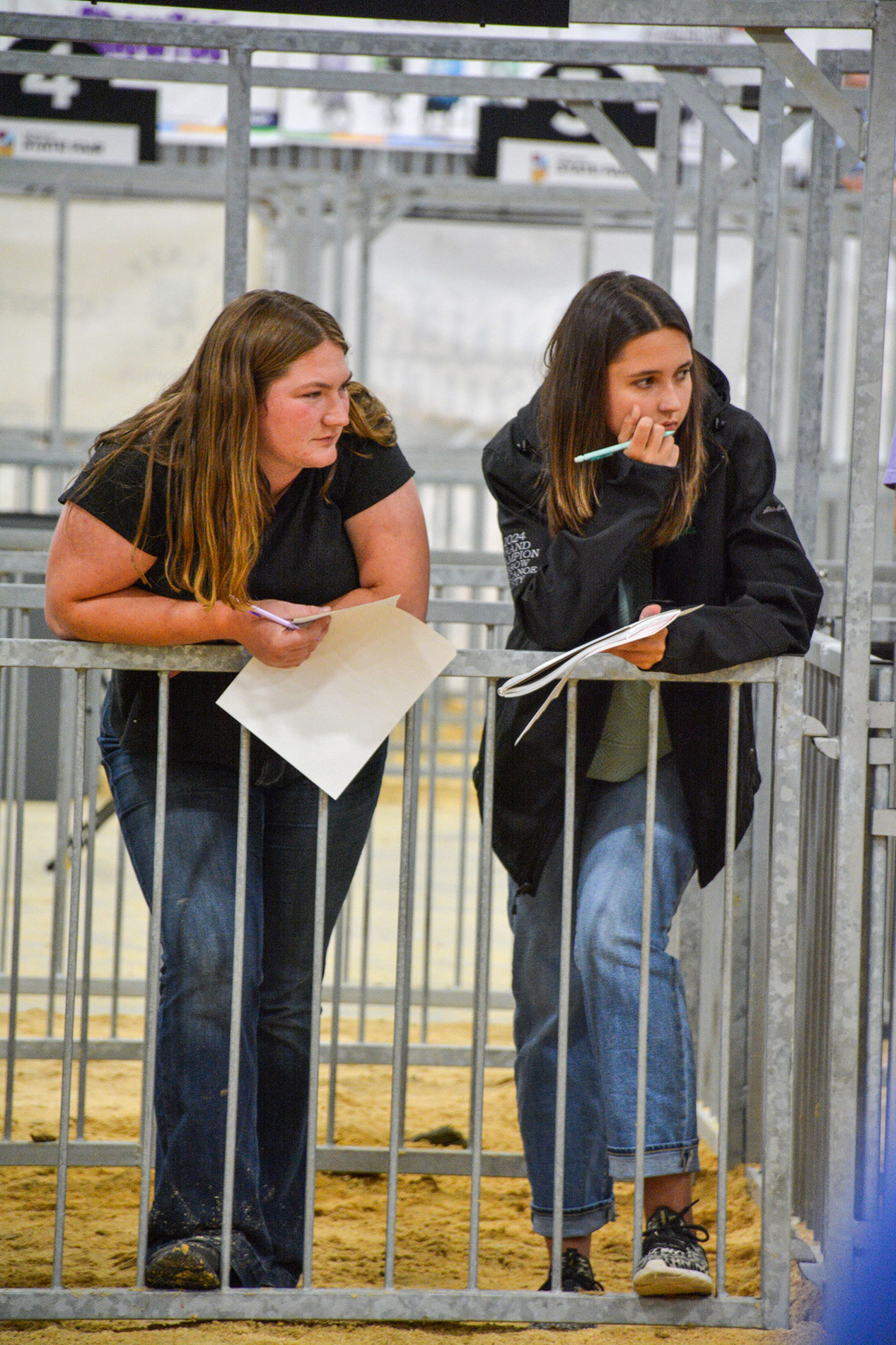Two individuals standing behind a metal railing in an indoor arena with a sandy floor, holding papers and writing utensils.