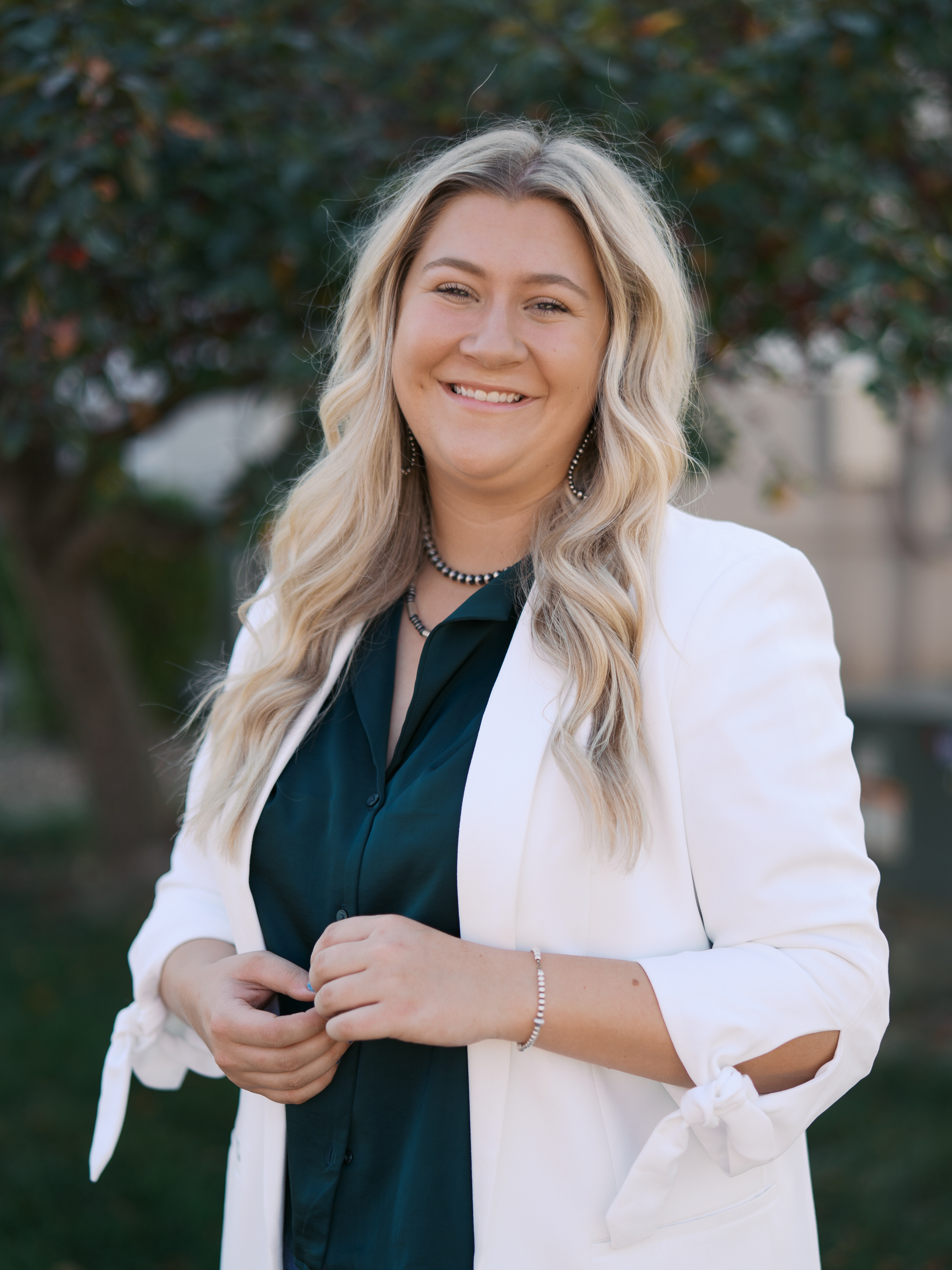 Student smiling outdoors, wearing a dark green blouse and white blazer, with trees softly blurred in the background.