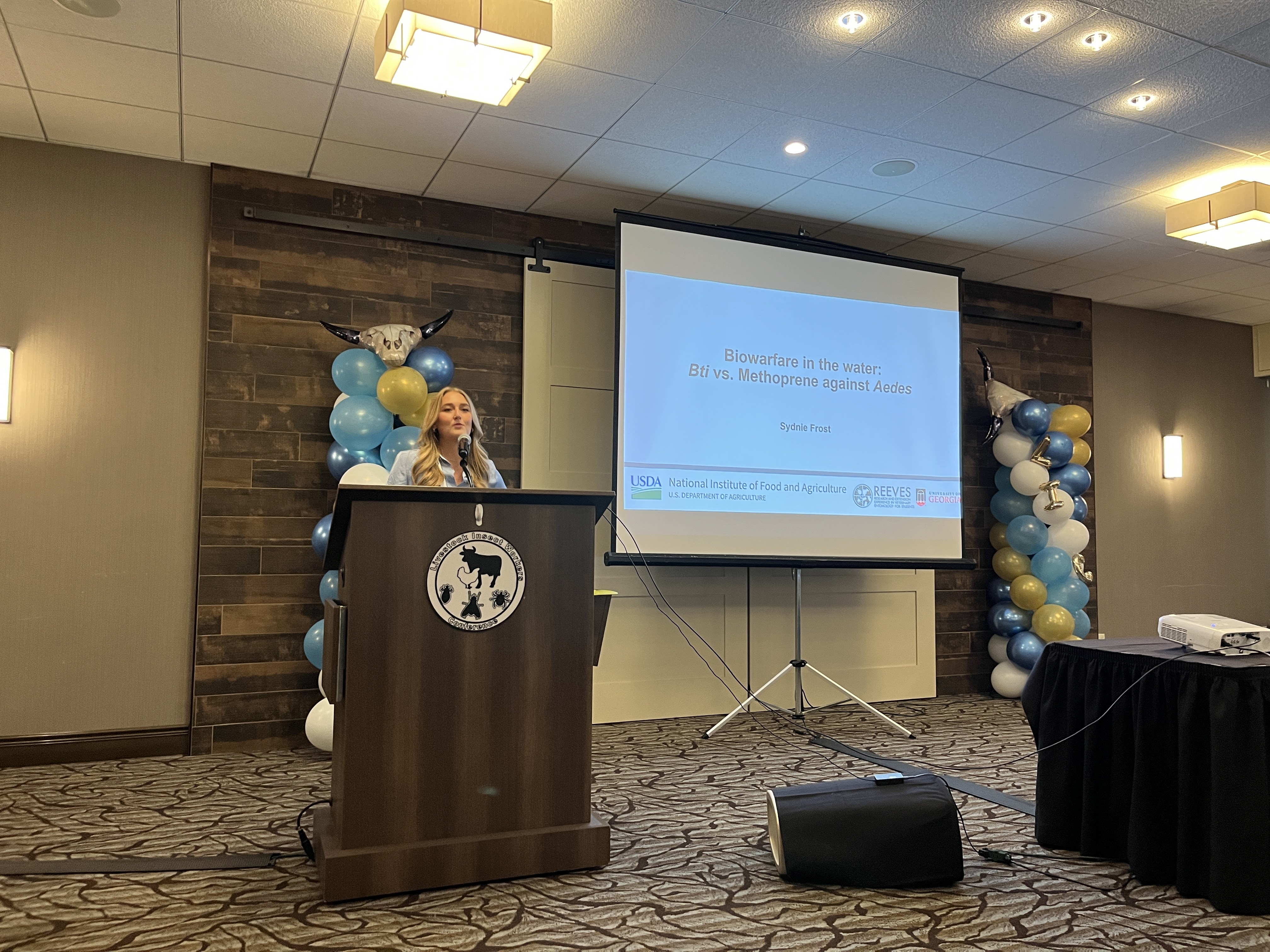 Student presenting research at a podium with a projected slide titled “Biowarfare in the water: Bti vs. Methoprene against Aedes,” with balloons and decorations in the background.