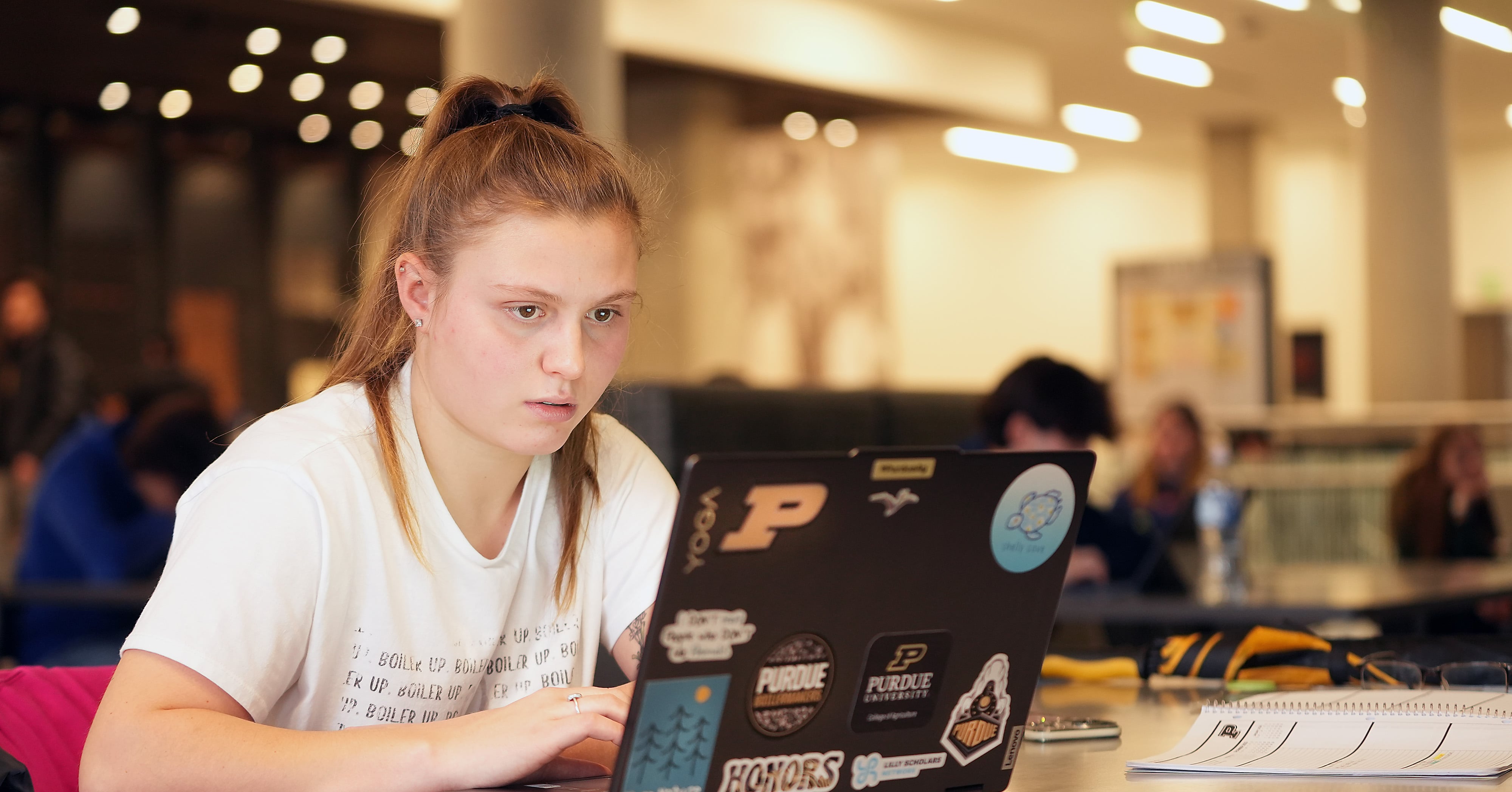 Student wearing a white T-shirt works on a laptop covered in Purdue stickers while sitting at a table with a notebook open. The background shows other students studying in a large, well-lit space.