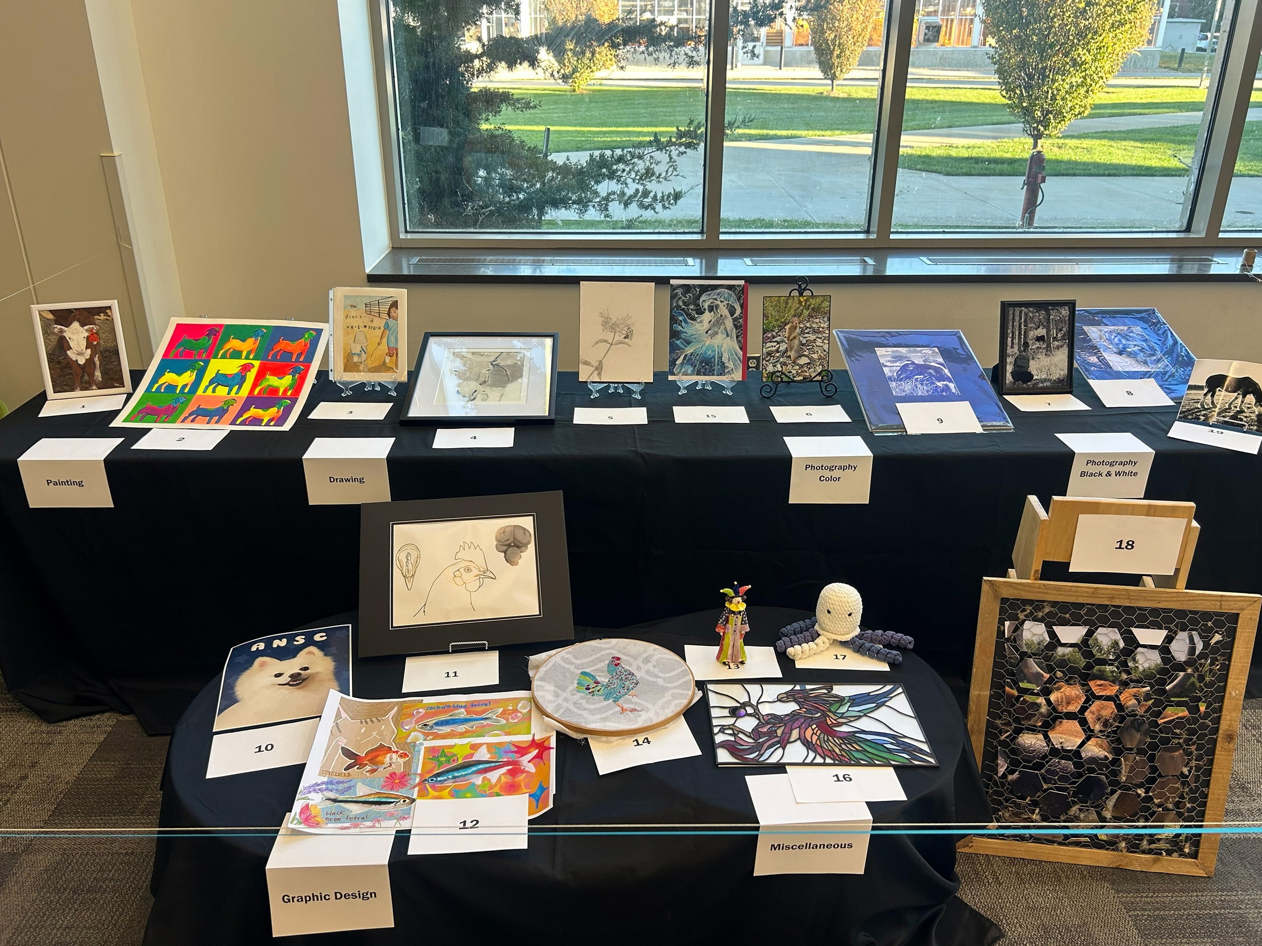 A display table in Creighton Hall features paintings, drawings, photographs, and mixed-media pieces from the Purdue Animal Sciences Art Show. Each artwork is labeled by category, including painting, drawing, photography, graphic design, and miscellaneous.