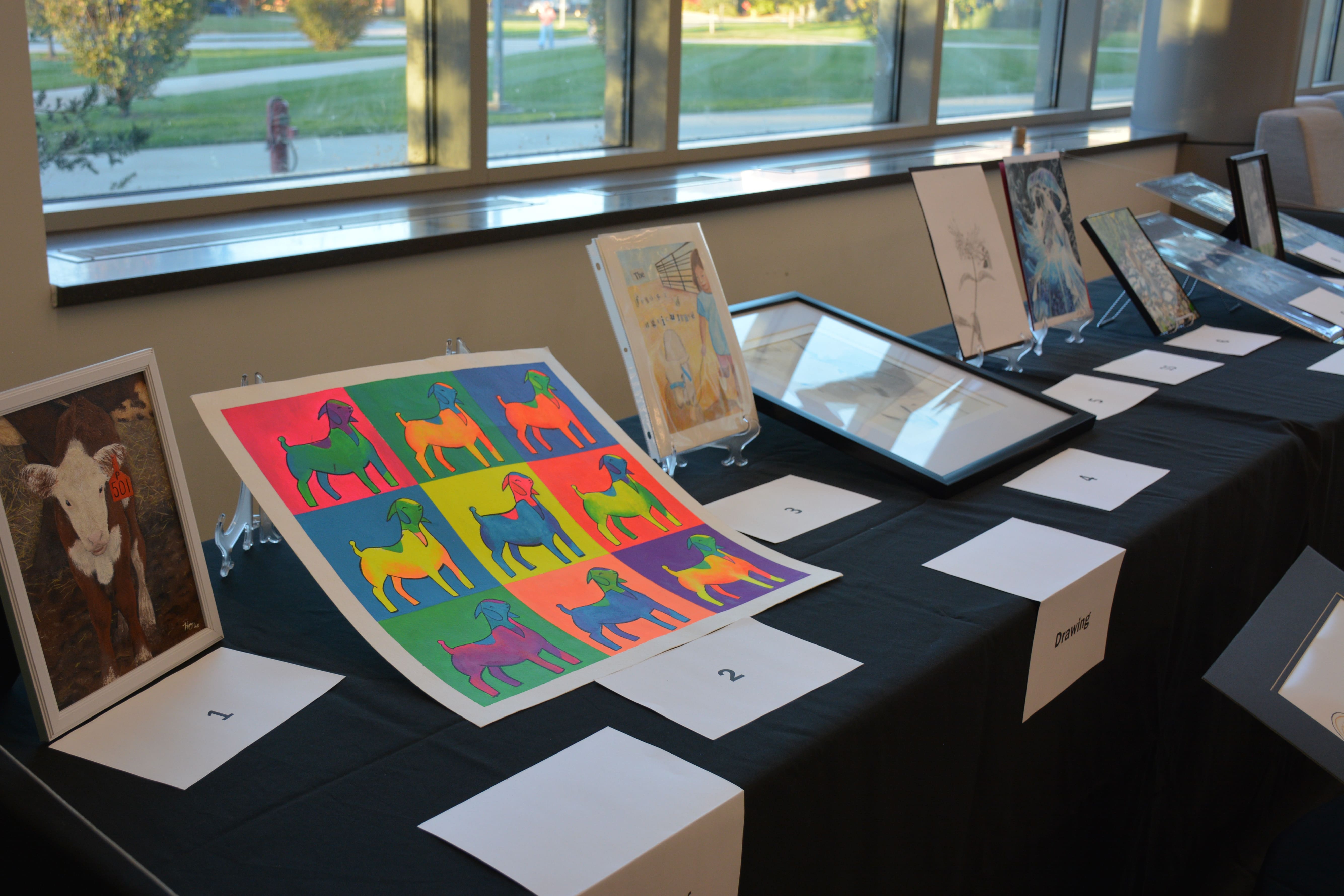 Framed student artwork from the Purdue Animal Sciences Art Show is displayed on black tablecloths near large windows in Creighton Hall. The pieces include colorful drawings, photography, and paintings labeled by category.