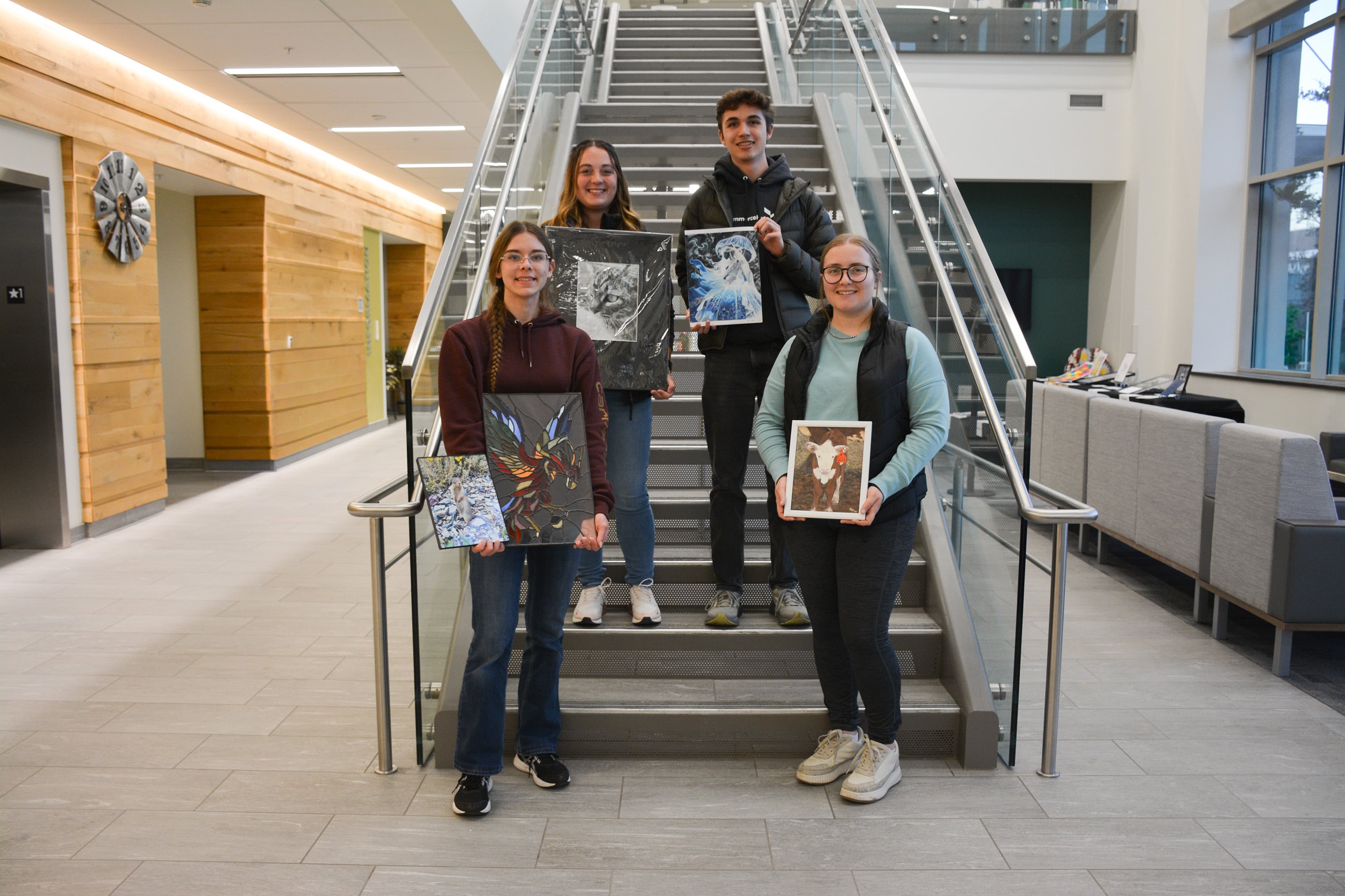 Four Purdue Animal Sciences students stand on the stairs inside Creighton Hall holding their winning art pieces from the department’s second annual art show. The photo shows colorful paintings and photographs as the students smile, celebrating their creativity and achievements.