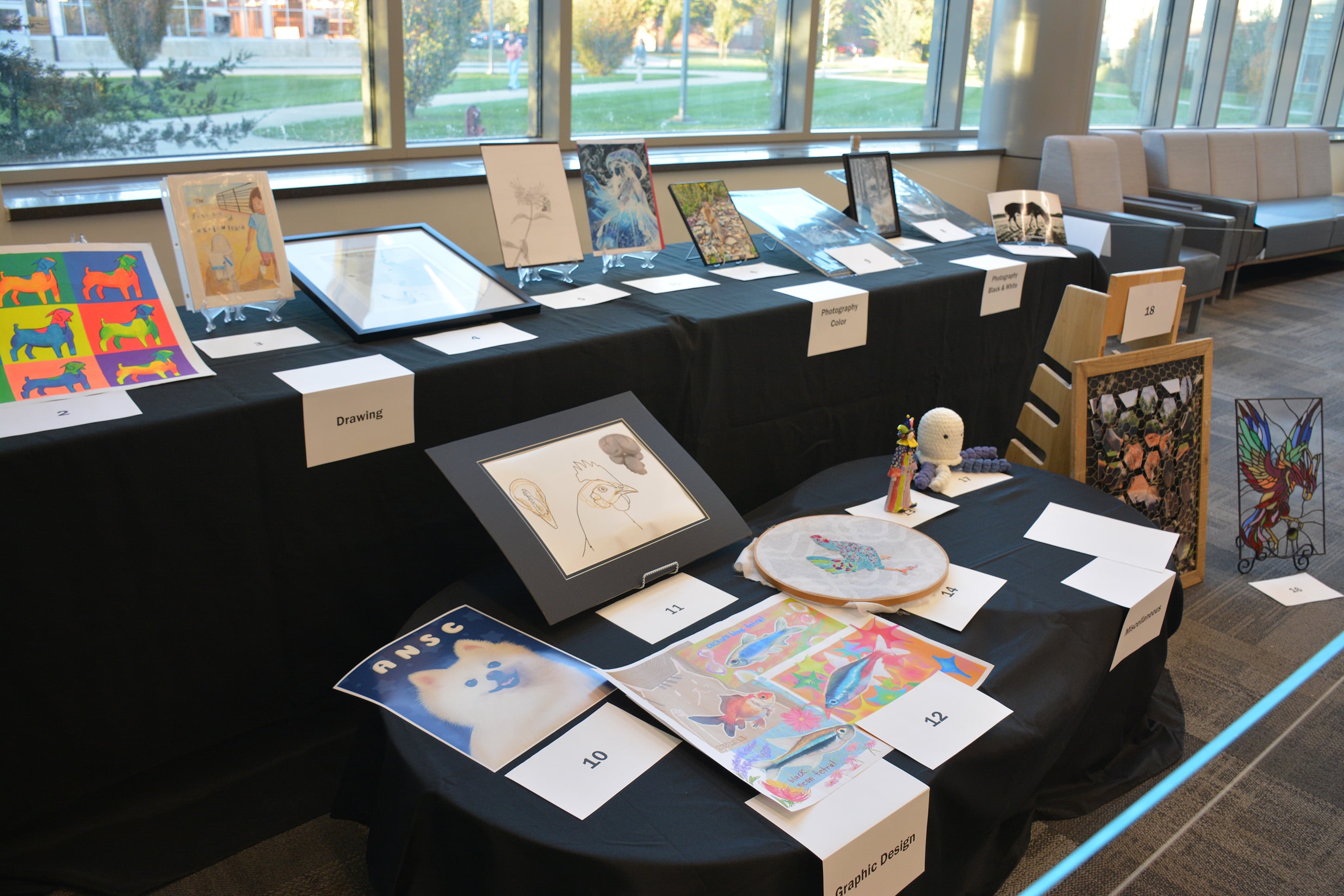 Artwork in the Graphic Design and Miscellaneous categories from the Purdue Animal Sciences Art Show is displayed on a table in Creighton Hall. Pieces include digital illustrations, embroidery, and a stained-glass–style creation.