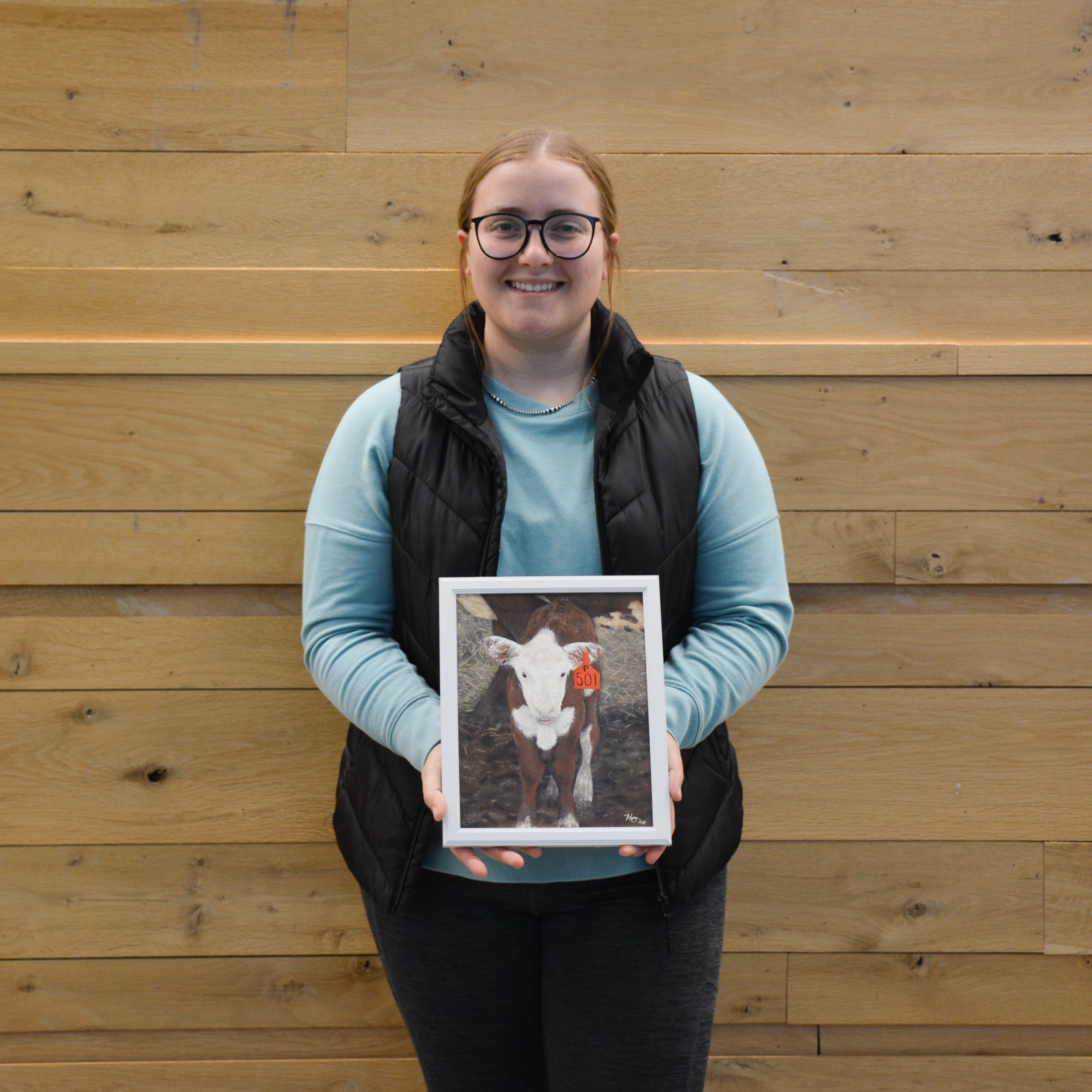 Hailey Orem smiles while holding her framed painting of a calf with a red ear tag. Her piece won the Painting category in the Purdue Animal Sciences Art Show. She stands in front of a light wood wall inside Creighton Hall.