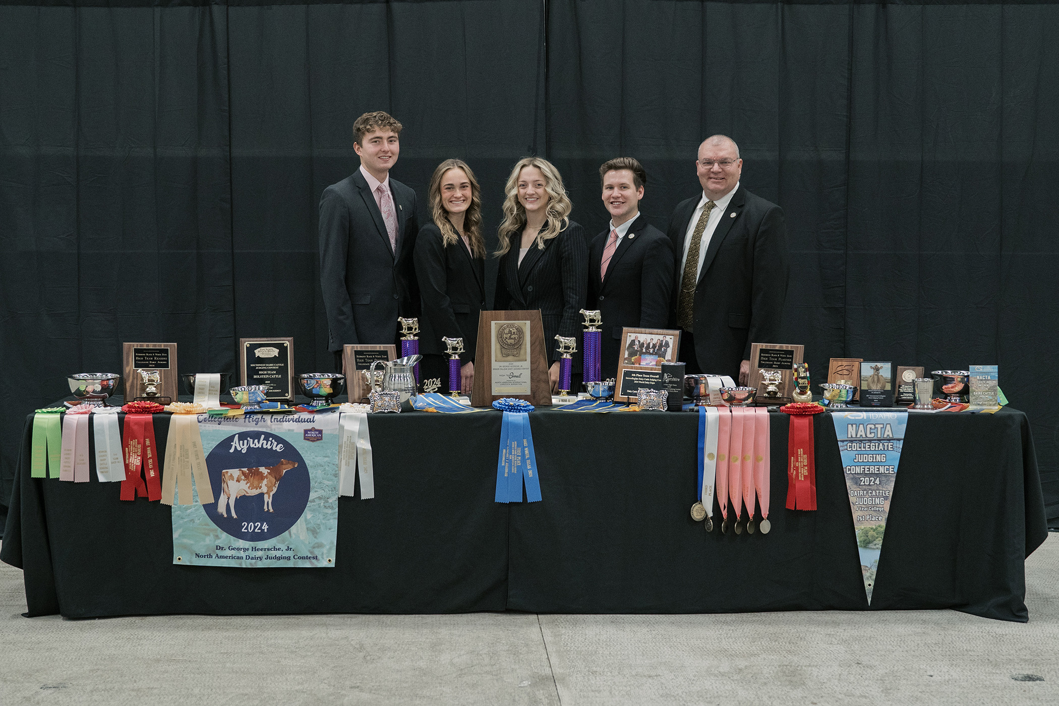 The Purdue Dairy Judging Team poses behind a table filled with trophies, ribbons, plaques and awards from multiple contests. The team stands side by side, dressed professionally in black suits.