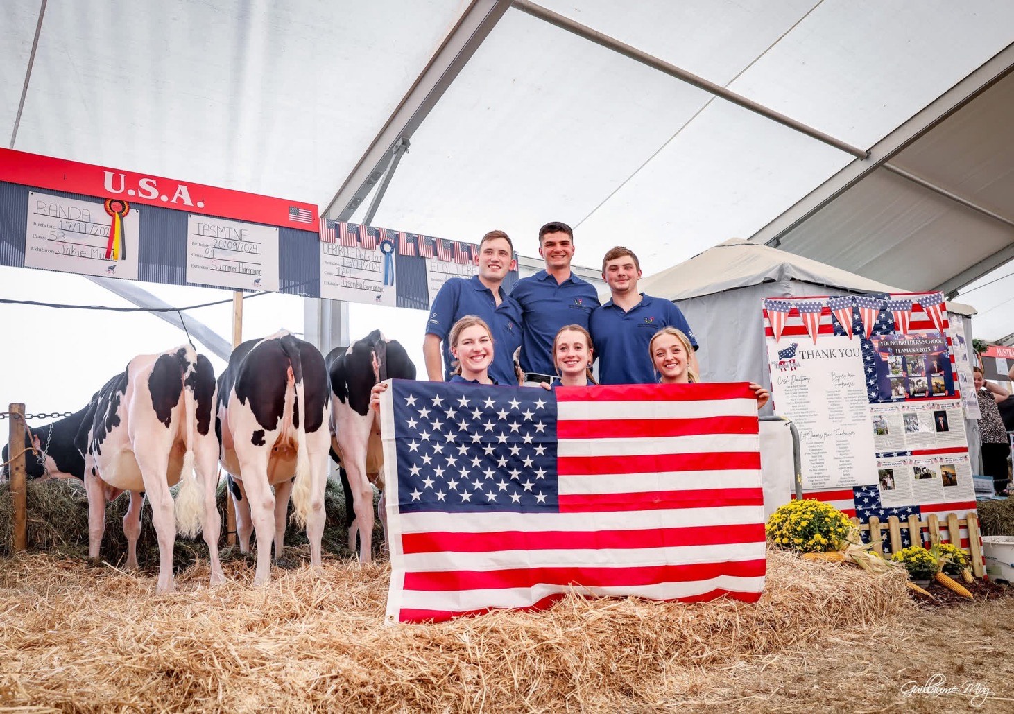 The U.S. team at the Young Breeders School poses in front of their cattle, holding an American flag. Jackie Mudd, wearing a blue shirt, is on the bottom row at the far right. The team stands in a decorated stall area with banners and signs displaying their names and sponsors.