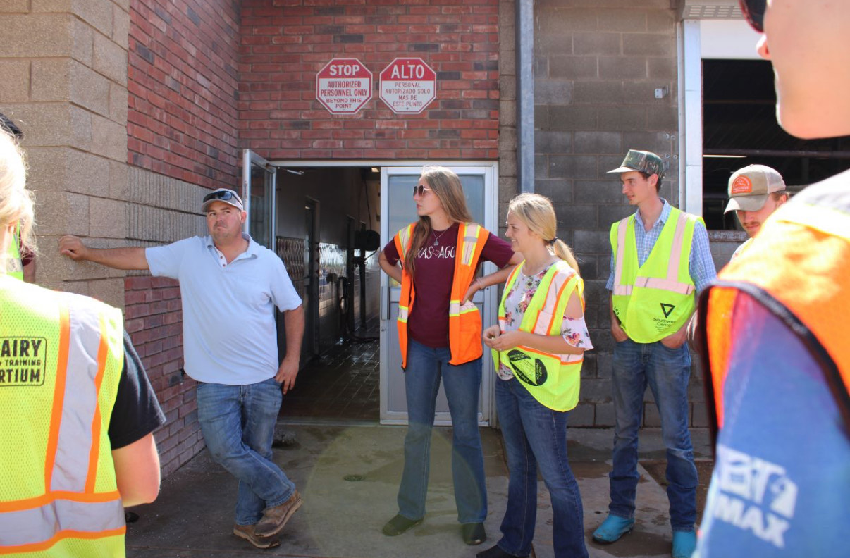 Jackie Mudd, wearing a floral shirt and yellow safety vest, stands outside a dairy facility listening to a manager speak during a tour. Other students in orange and yellow vests stand nearby under a sign reading “Authorized Personnel Only.”