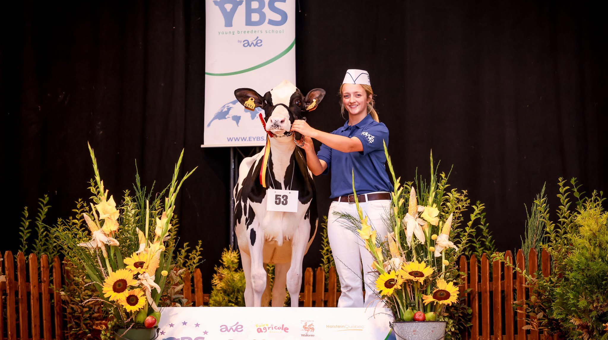 Jackie Mudd stands beside a Holstein cow wearing a numbered tag at the Young Breeders School in Belgium. She’s smiling and holding the cow’s halter in front of a stage decorated with sunflowers, greenery and event banners.