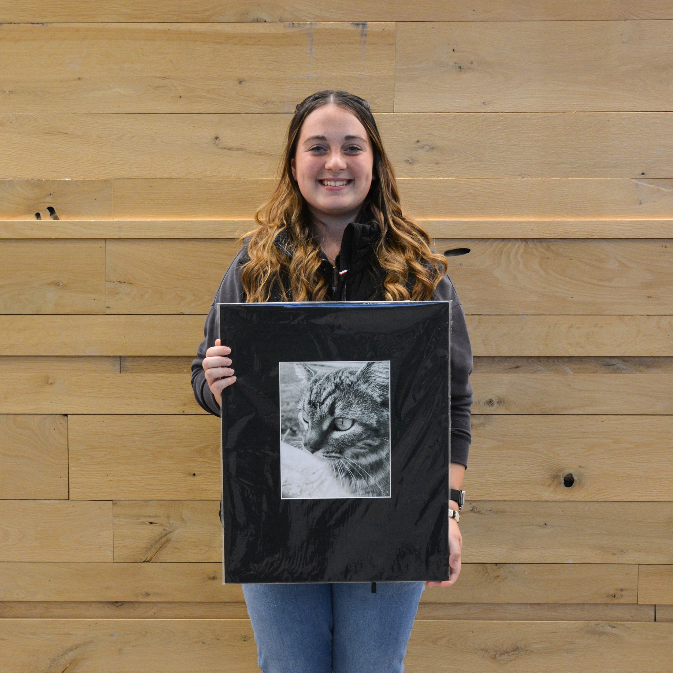 Kara Baker holds her black-and-white photograph of a cat, which earned first place in the Black and White Photography category of the Purdue Animal Sciences Art Show. She stands in front of a light wood wall inside Creighton Hall, smiling for the photo.