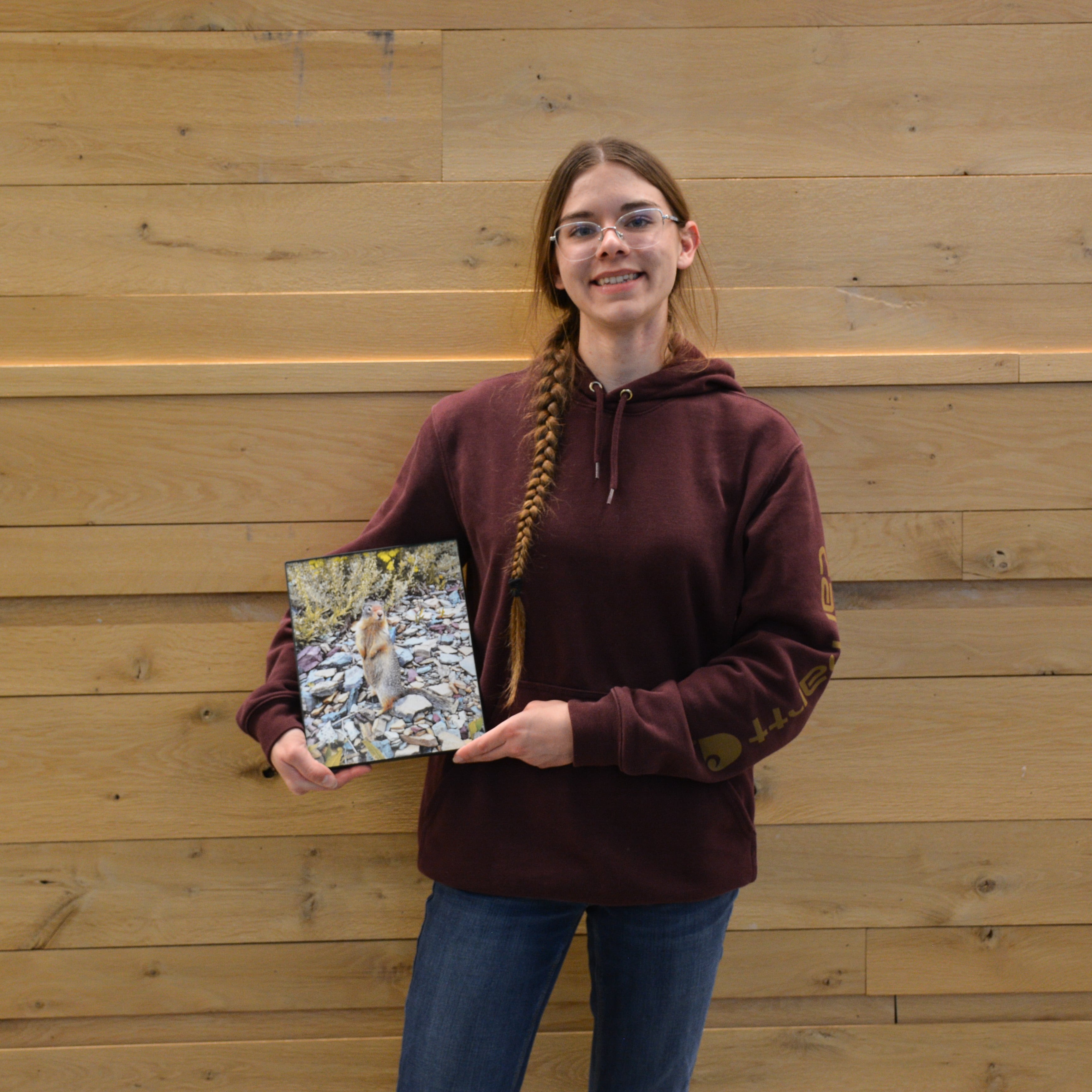 Sarah Tepp holds her color photograph of a squirrel standing among rocks, which earned her first place in the Color Photography category of the Purdue Animal Sciences Art Show. She stands in front of a light wood wall inside Creighton Hall.