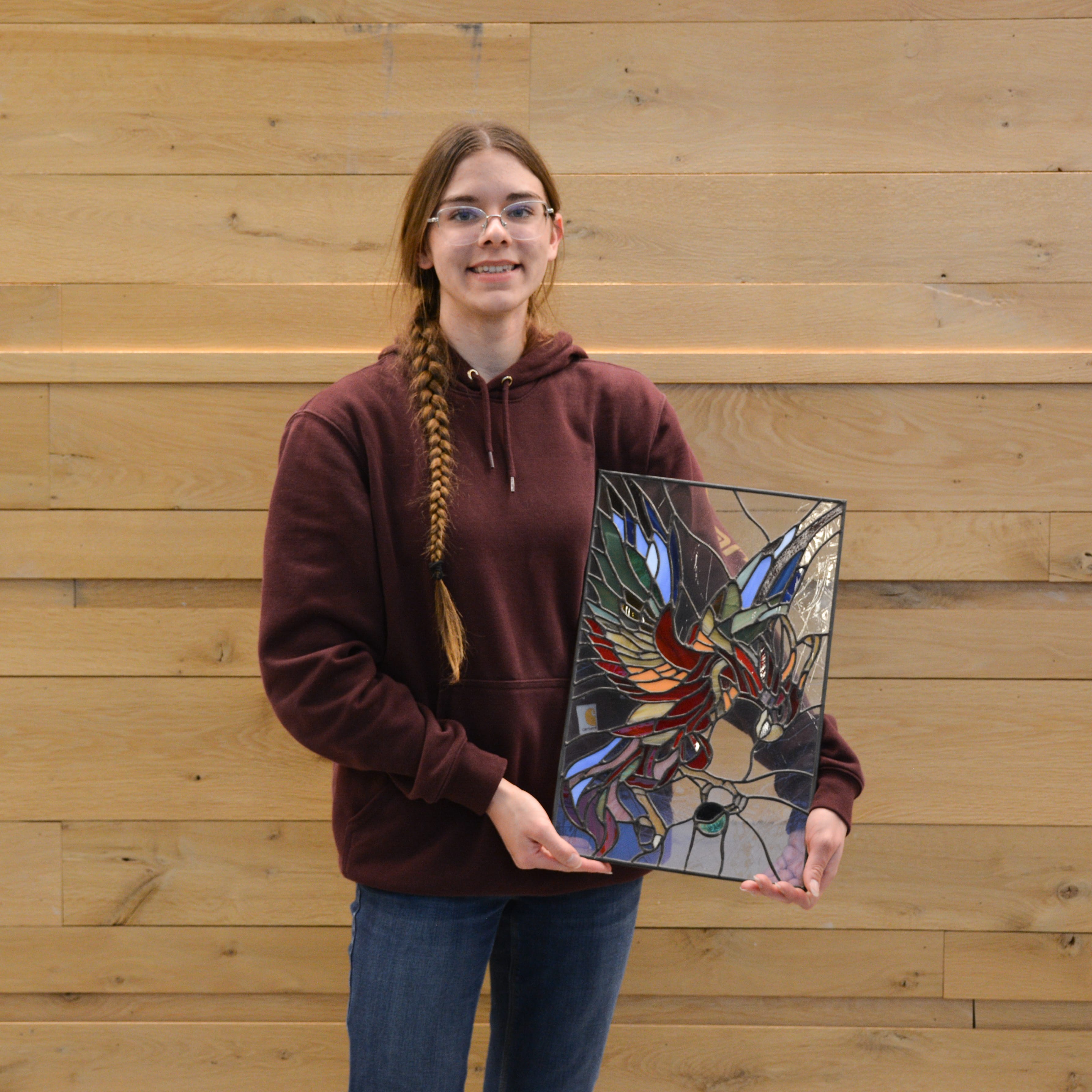 Sarah Tepp holds her colorful mixed-media artwork, which won the Miscellaneous category in the Purdue Animal Sciences Art Show. She stands in front of a light wood wall inside Creighton Hall, smiling for the photo.
