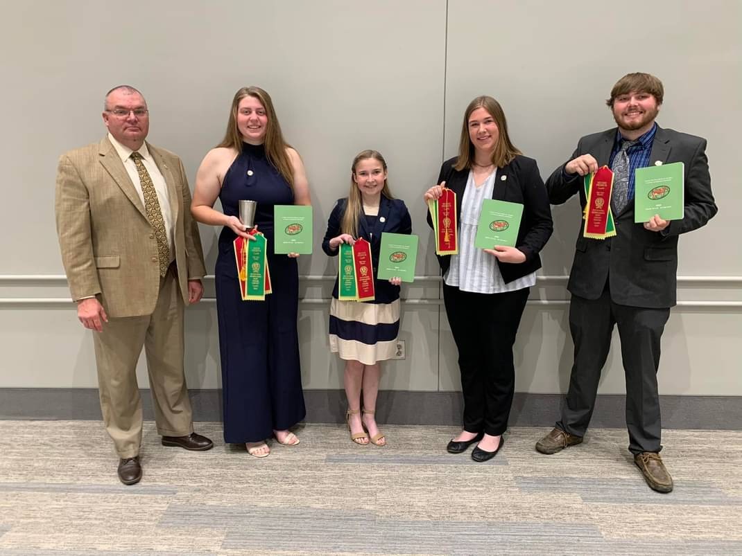 Brian Engleking and the 2023 Purdue Dairy Judging Team pose indoors holding green award books and red and yellow ribbons after a contest.