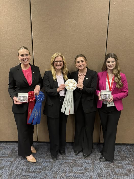 Four members of the Purdue Dairy Judging Team stand in front of a tan wall, smiling and holding ribbons and plaques from a competition.