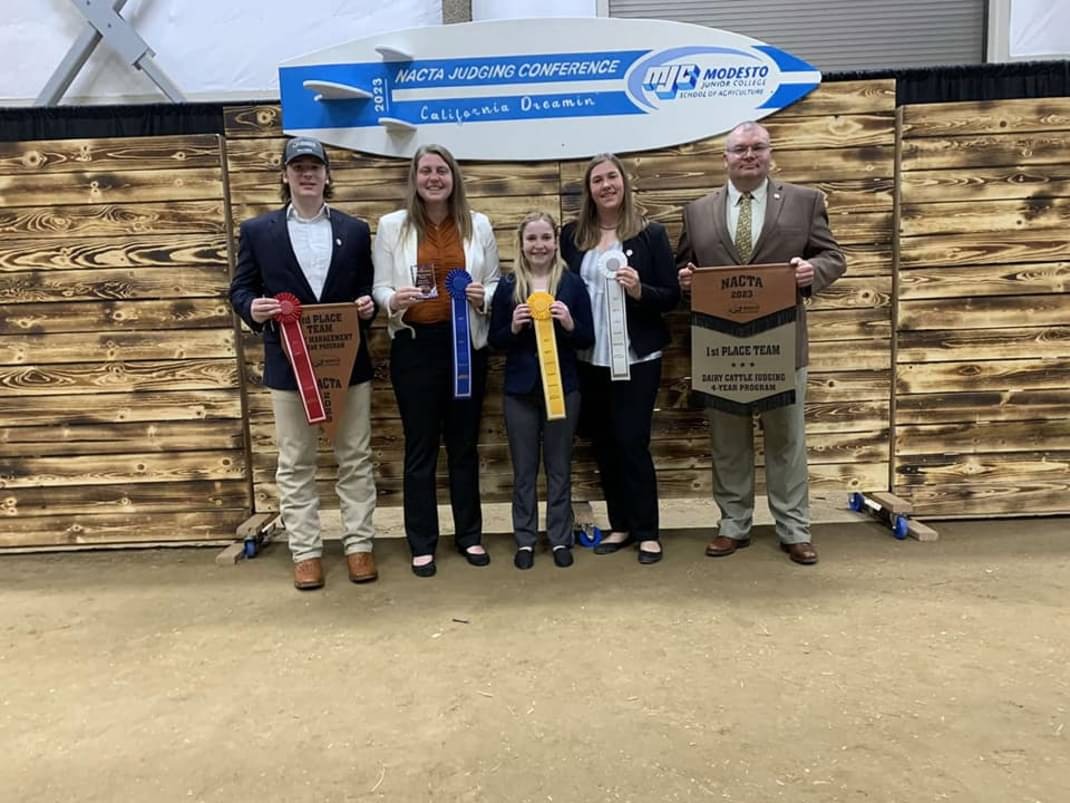 The 2023 Purdue Dairy Judging Team, including coach Brian Engleking, stands in front of a wooden backdrop holding ribbons and banners after winning first place in the dairy cattle judging contest at the NACTA Judging Conference.