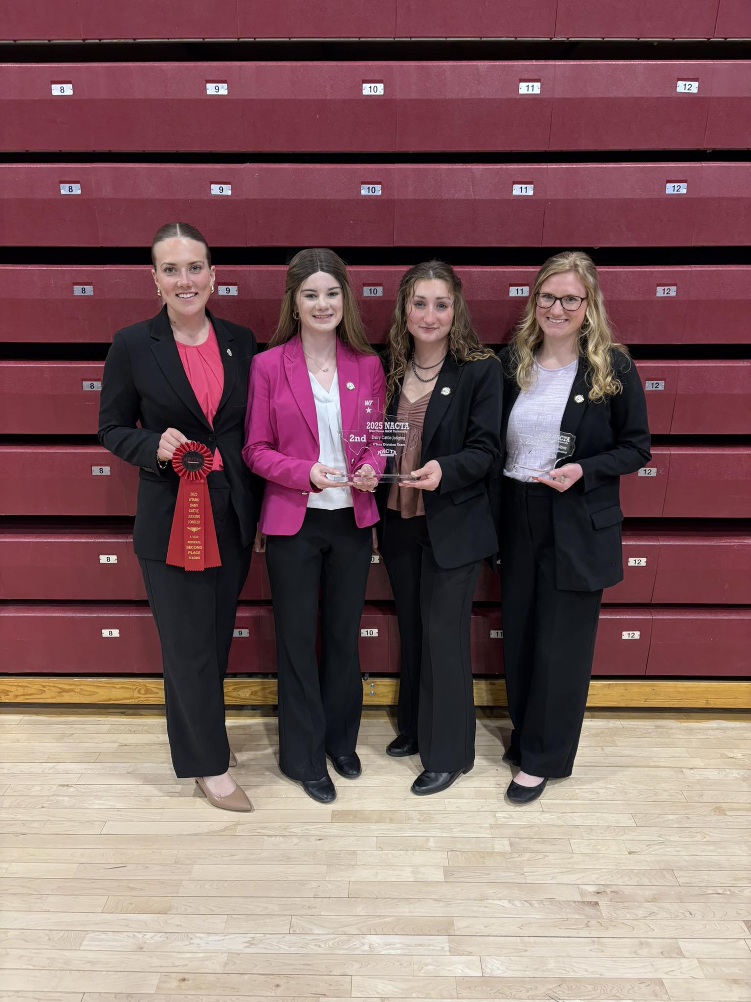 Four members of the Purdue Dairy Judging Team stand in front of red bleachers, each holding awards and ribbons earned during the 2025 NACTA Judging Conference.
