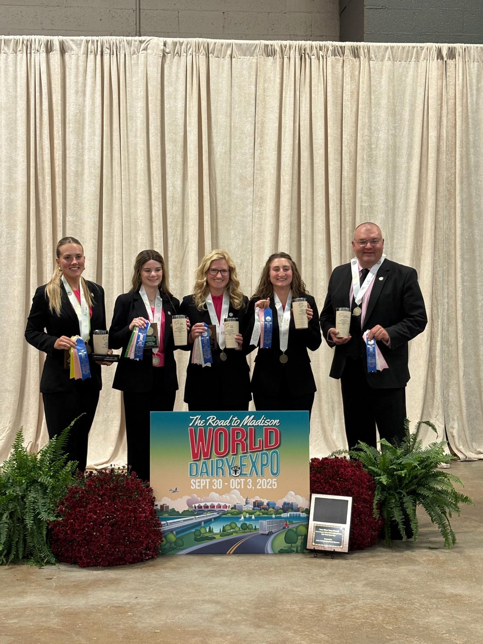 The Purdue Dairy Judging Team stands together indoors in front of a beige curtain, holding ribbons and awards from the 2025 World Dairy Expo. Four female team members and coach Brian Engleking, all dressed in black suits, smile proudly behind a colorful World Dairy Expo sign.