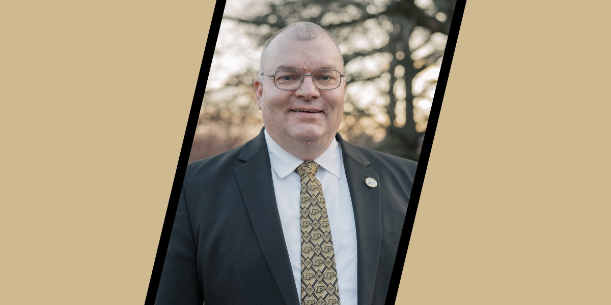Brian Engleking, wearing glasses, a black suit jacket, and a white shirt with a gold Purdue-patterned tie, smiles while standing outdoors with trees softly blurred in the background.