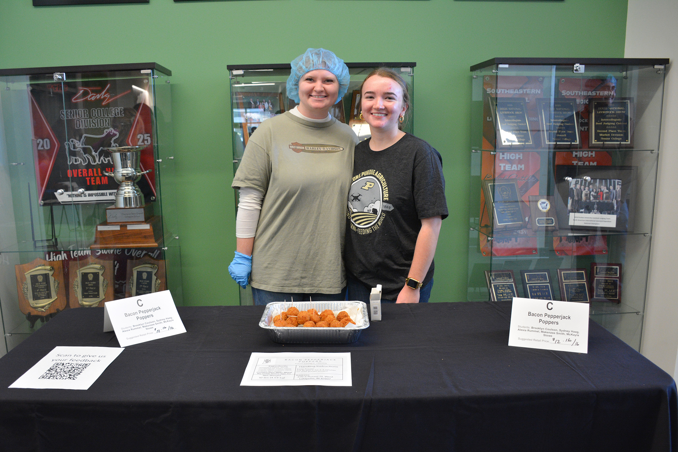 Students stand behind a display table presenting their Bacon Pepperjack Poppers, with trays of fried poppers and product signage at the ANSC 360 Product Show.
