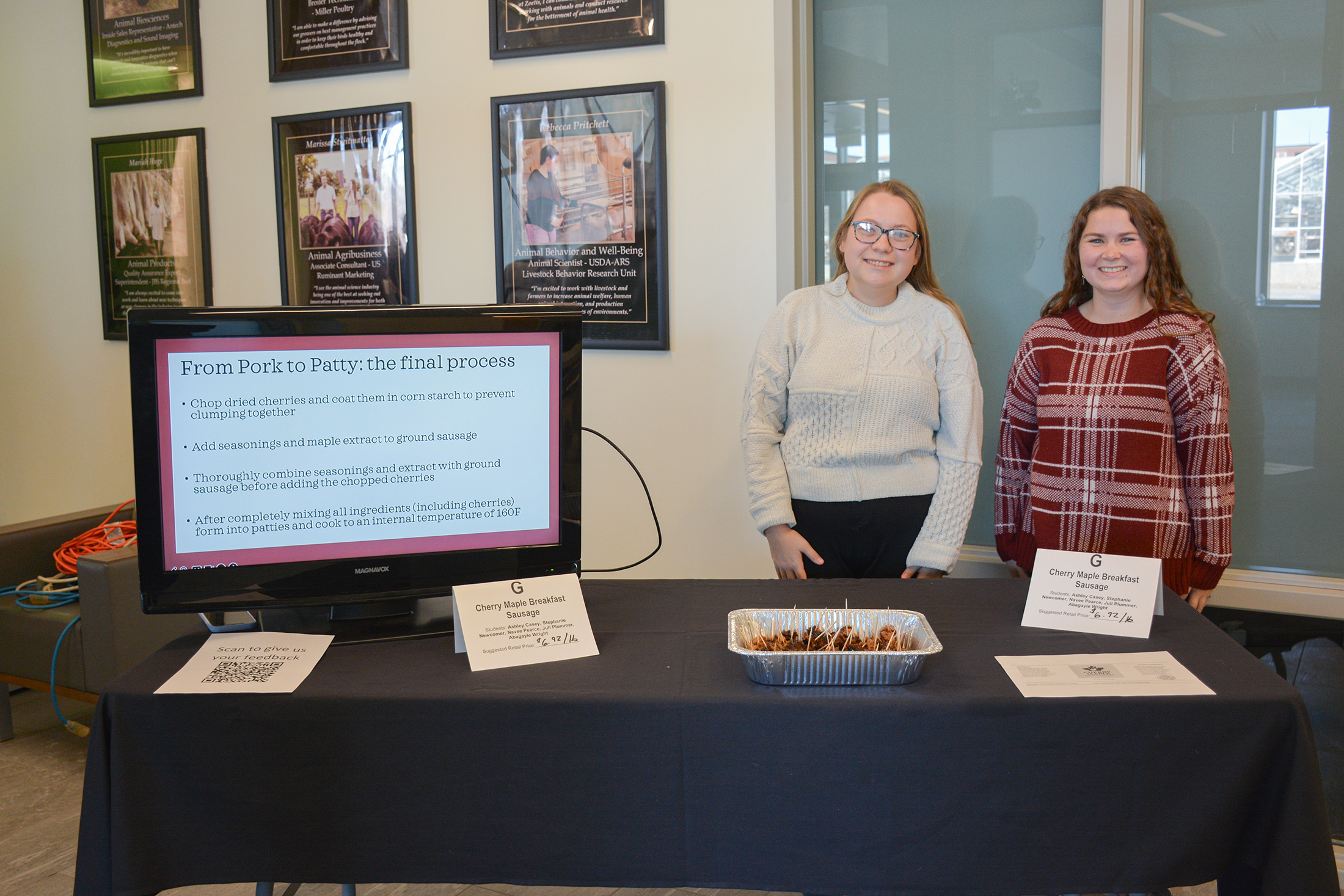 Students stand behind a display table presenting their Cherry Maple Breakfast Sausage, with sample trays and a screen explaining the product process at the ANSC 360 Product Show.