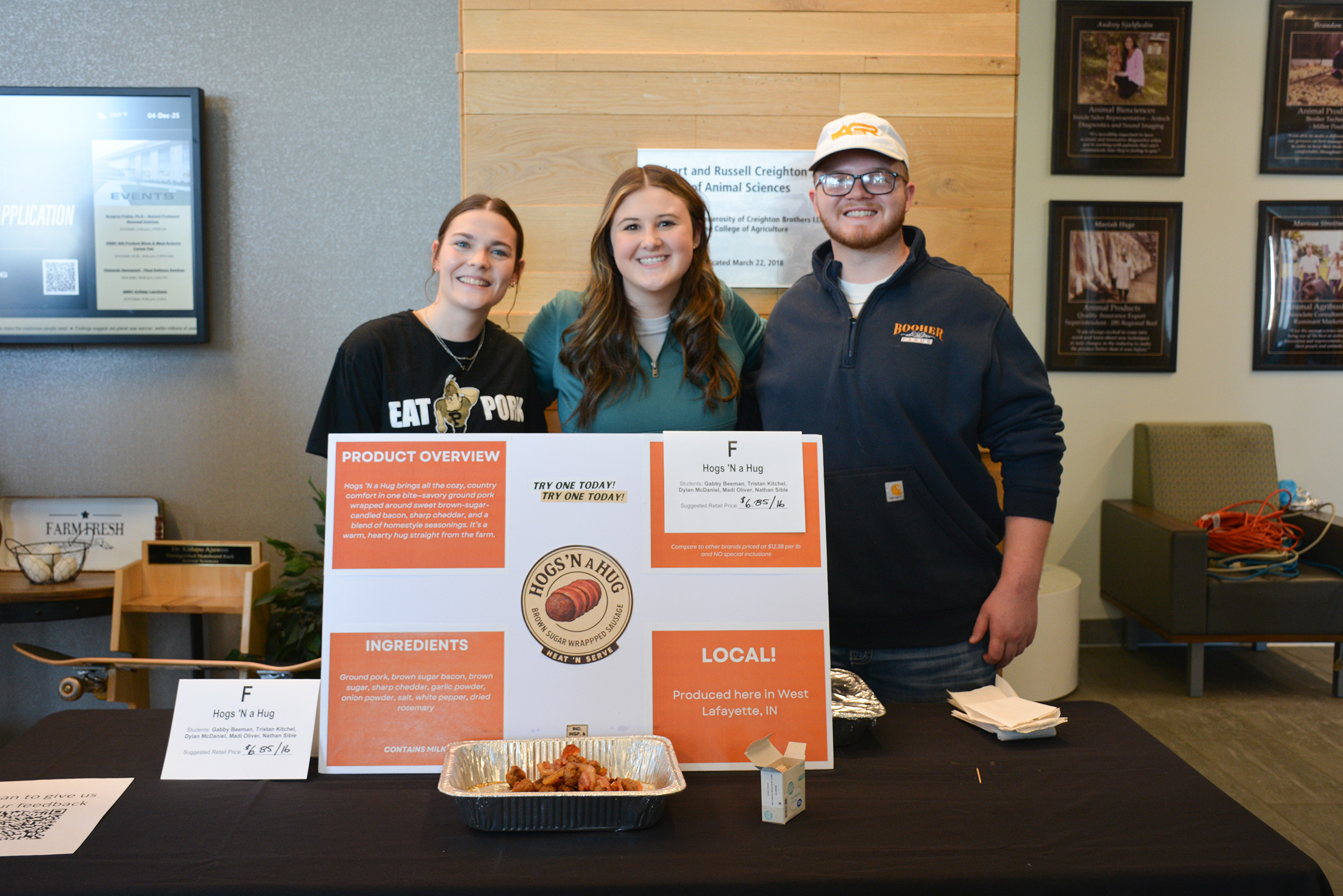 Students stand behind a display table presenting their Hogs ’N a Hug product, with signage and sample trays of ground pork wrapped in candied bacon at the ANSC 360 Product Show.