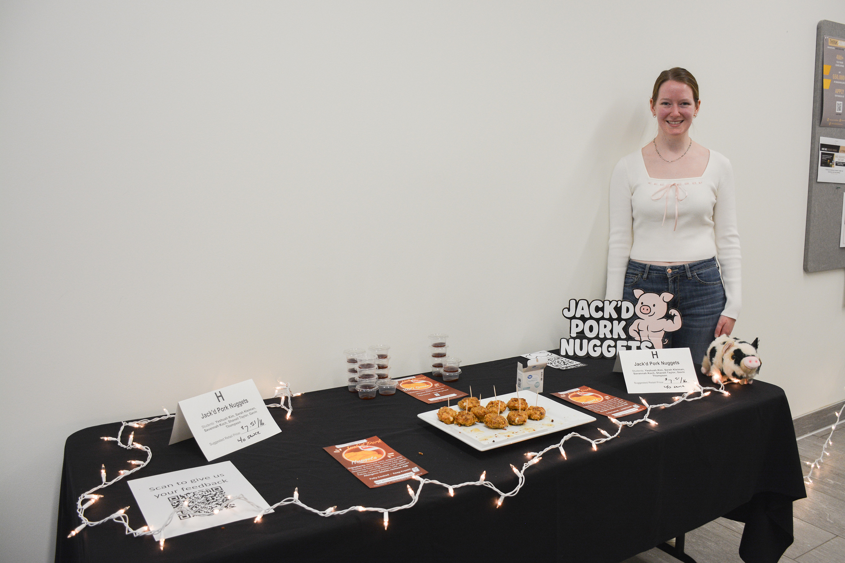A student stands behind a display table presenting Jack’d Pork Nuggets, with plated samples, product signage and promotional materials at the ANSC 360 Product Show.