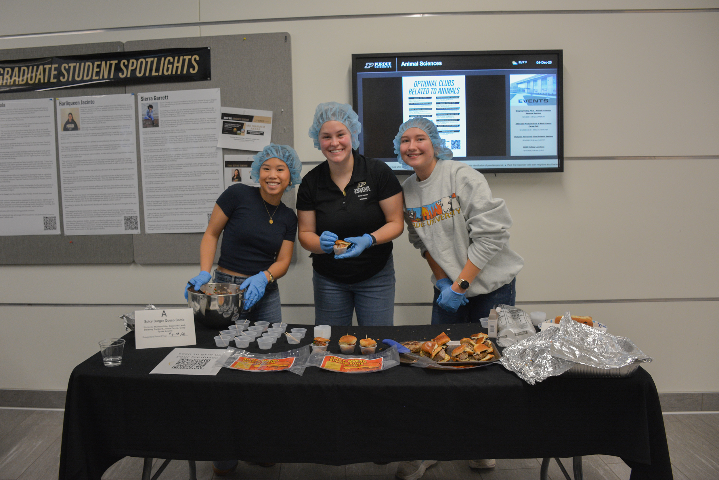 Students wearing hairnets and gloves stand behind a table serving samples of their Spicy Burger Queso Bomb during the ANSC 360 Product Show.