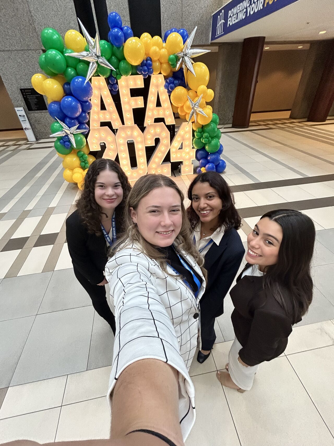 Four students smiling for a selfie inside a convention center, standing in front of a colorful balloon display with illuminated letters reading “AFA 2024.”