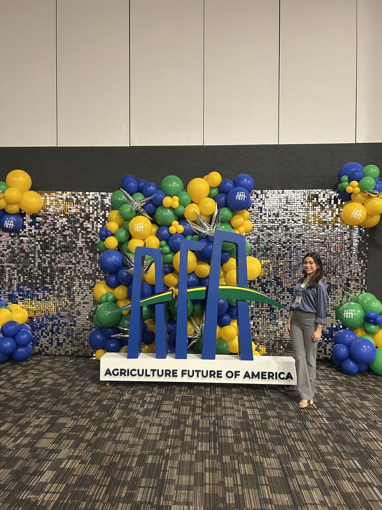 A student stands smiling beside a large Agriculture Future of America (AFA) logo surrounded by blue, green and yellow balloon displays inside a conference venue.