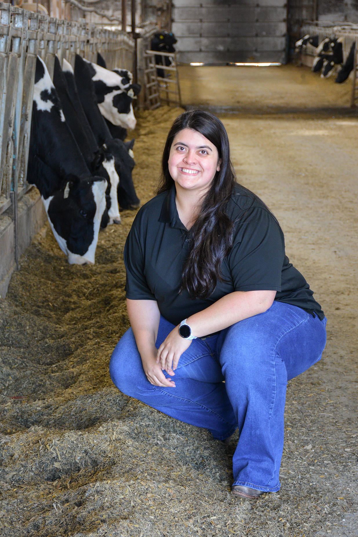 Isabela Santos kneels in a dairy barn aisle with cows eating behind her.