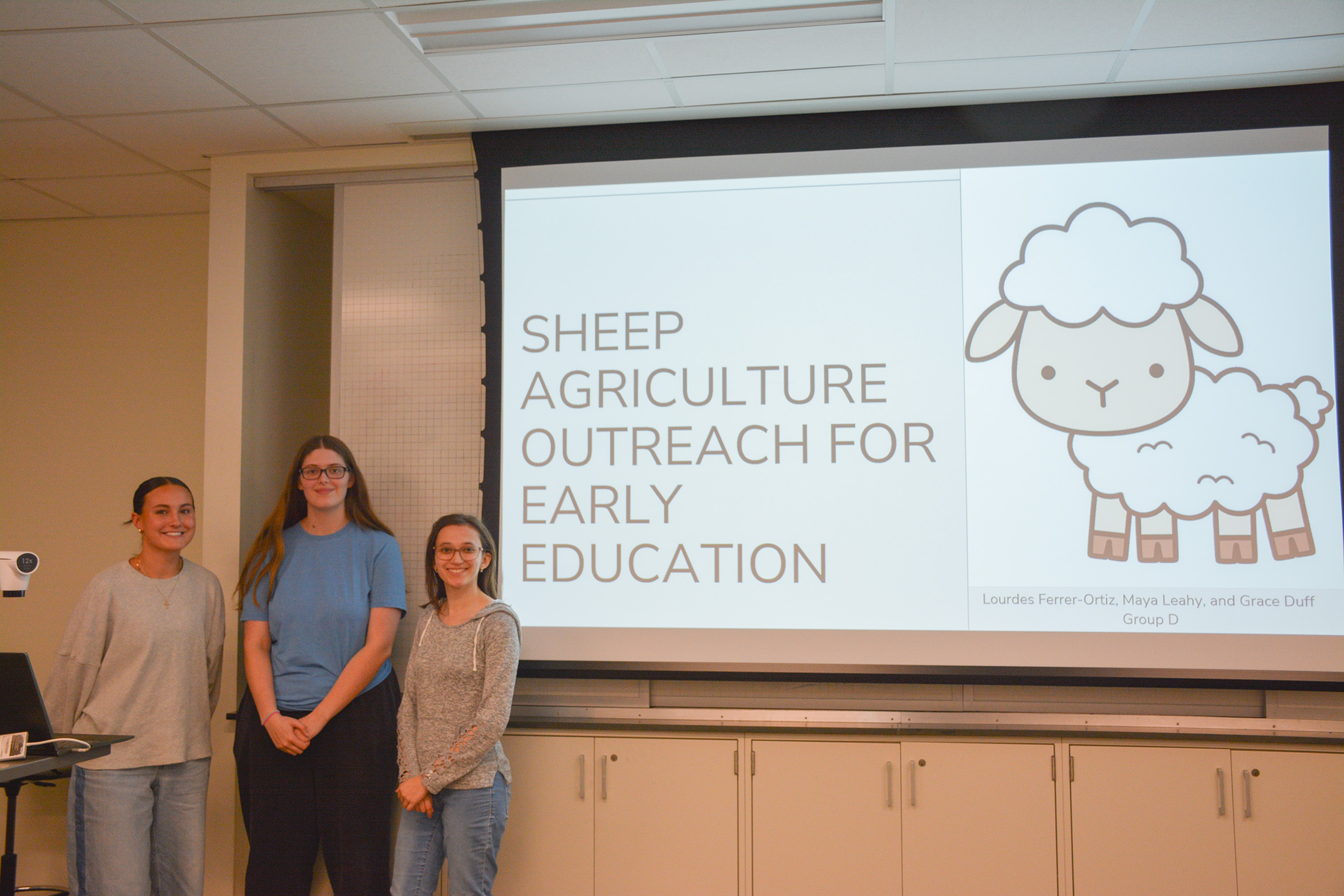 Students stand in front of a presentation that's titled "Sheep Agriculture Outreach for Early Education."