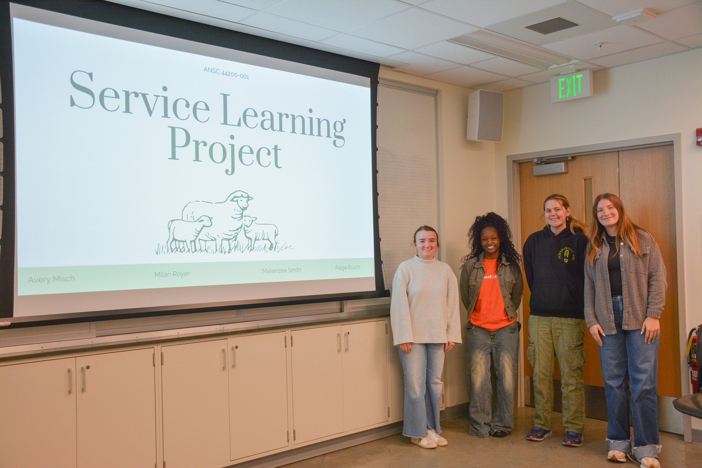 Students stand in front of a presentation that's titled "Service Learning Project."