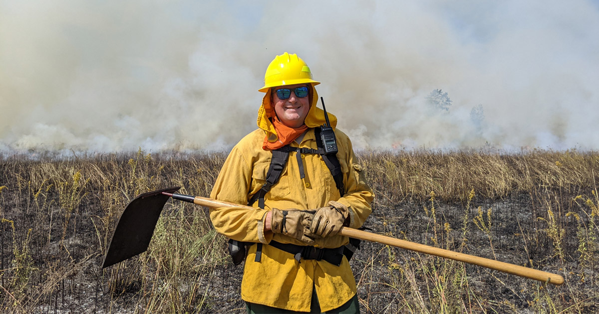 Jarred Brooke holds a flapper in front a field that is smoking after a prescribed burn.