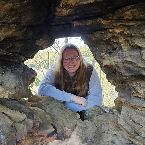 Alex Dudley holds a black vulture; Alex is pictured through a hole in a rock formation; Alex holds her camera in front of a forested mountain landscape. 