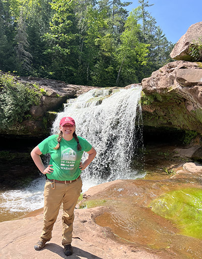 Alex Dudley stands in front of a waterfall and a forested hillside