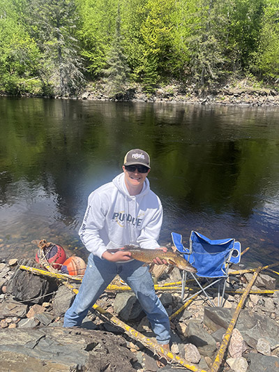 Austin Grant holds a fish in front of a body of water and forest area.