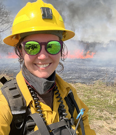 Danielle Howard stands in front of a field on fire from a prescribed burn.