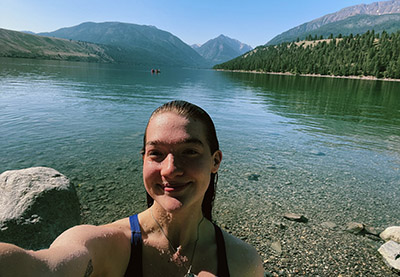 Alyssa Johnson is pictured in front of a water body with a mountainous and forested landscape behind her.