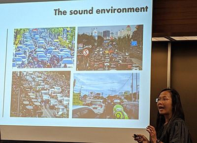 Dr. Christina Mediastika stands next to a slide showing various noises sources in Indonesia in the form of traffic
