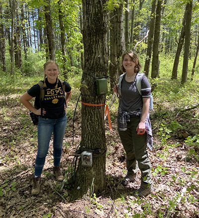 Alexis Proudman and co-author Morgan Watkins pose in front of a trail camera and acoustic monitor during their 2021 cicada research